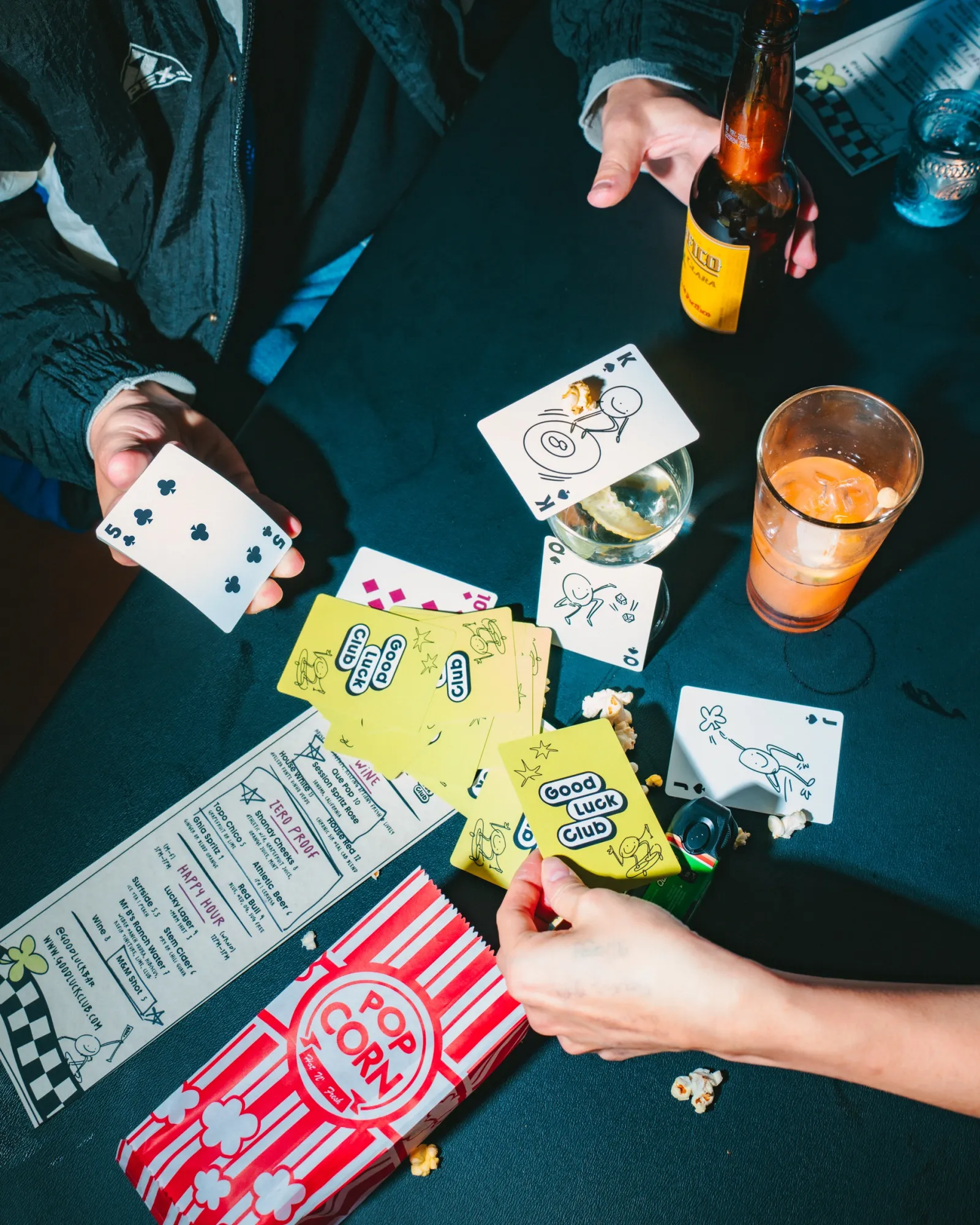 Overhead shot of hands playing cards around a table with drinks, the menu and a popcorn box