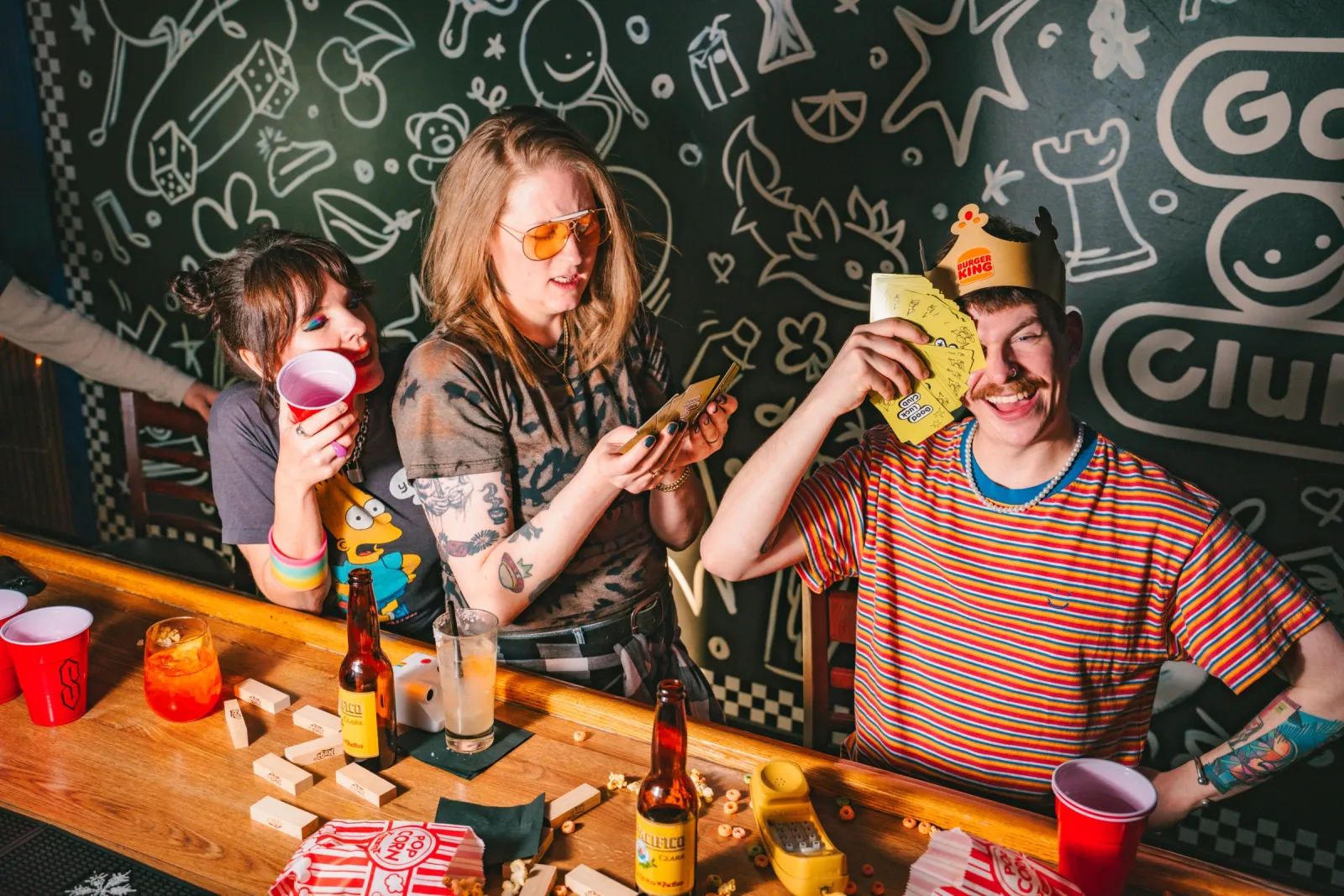 Friends playing a card game at the bar under the doodle mural, red cups and popcorn on the bar