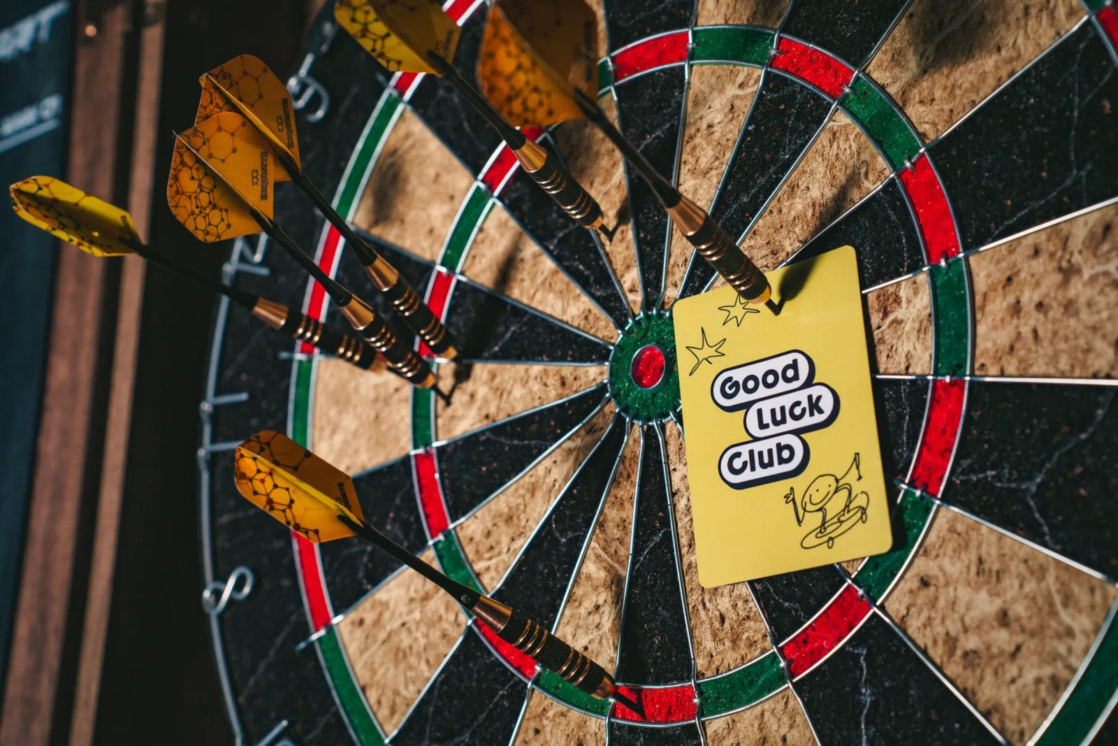 Darts stuck in a dartboard with a Good Luck Club card pinned to it
