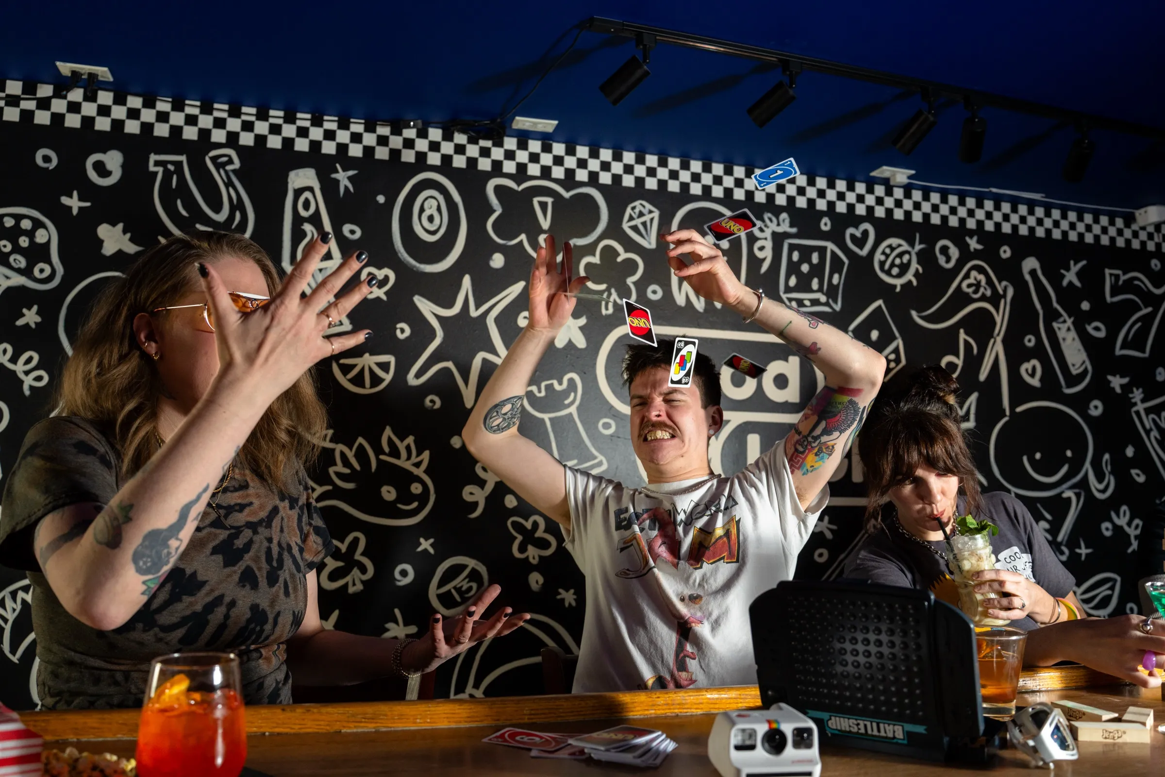 Friends at the bar in front of the black-and-white doodle mural, mid-game, arms in the air