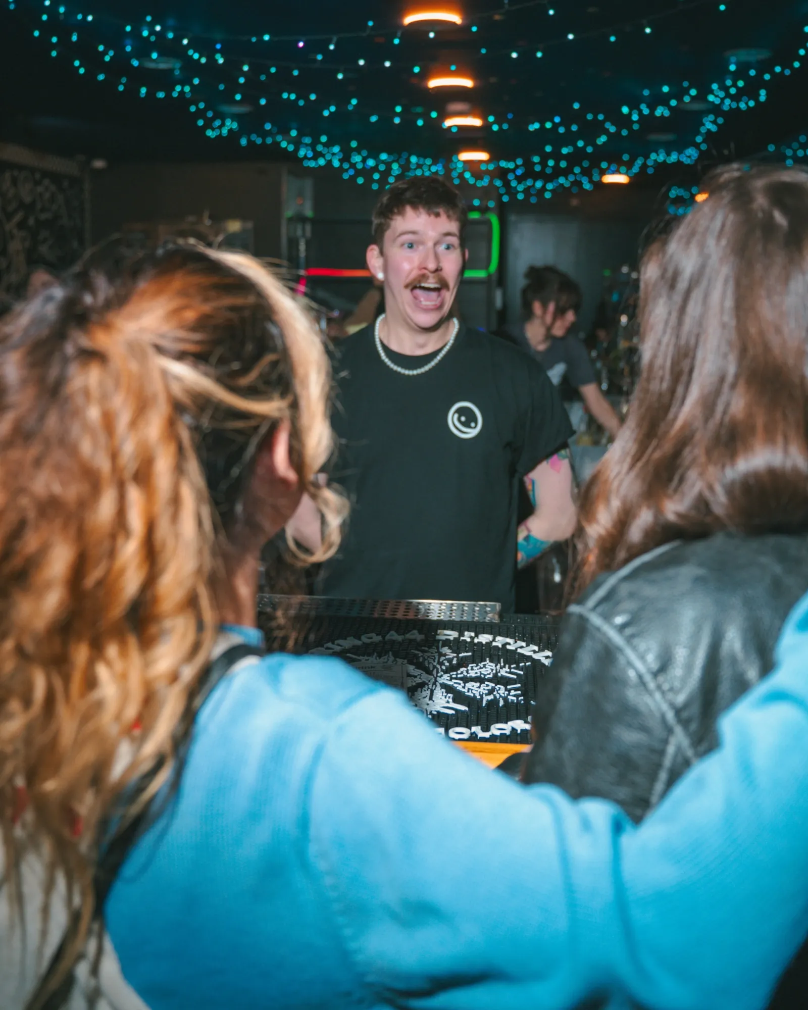 A bartender laughing with two guests across the bar under blue string lights