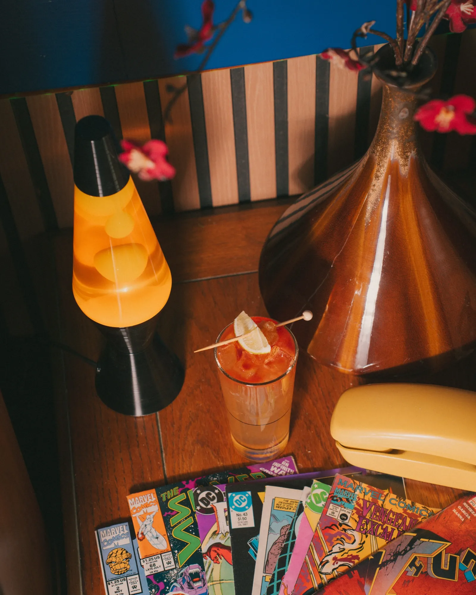A cocktail beside a lava lamp, a vintage vase and a yellow phone, comic books on the table