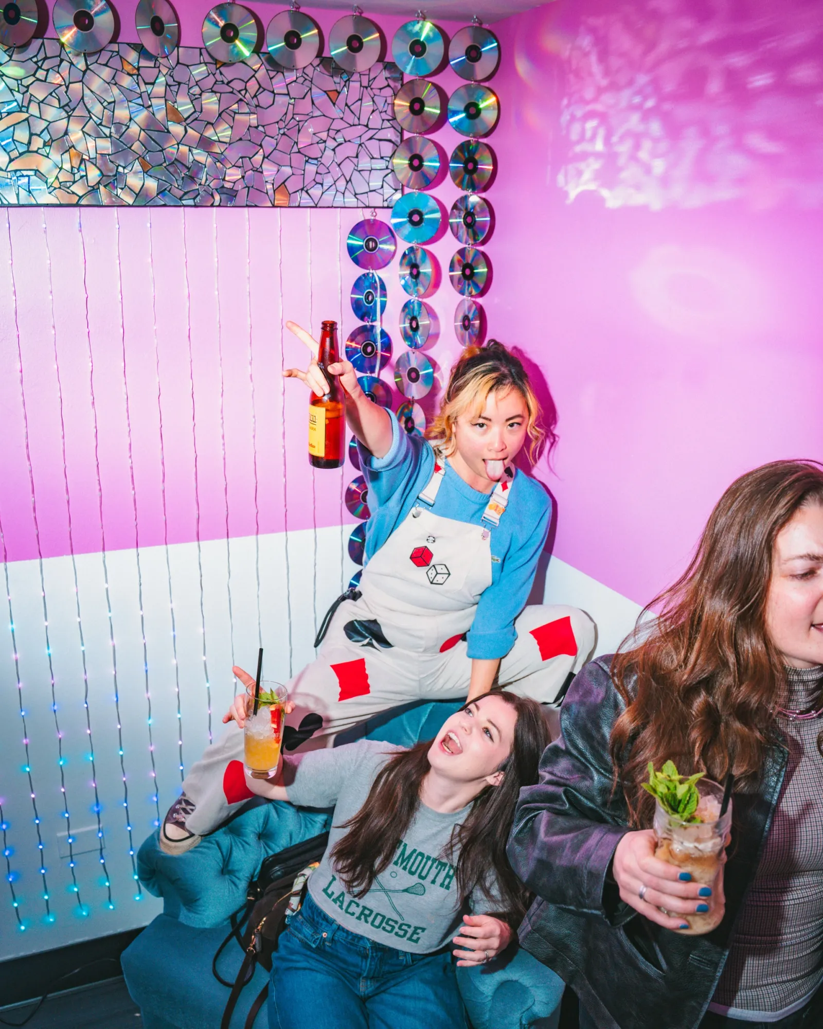 Friends laughing on the teal couch in the pink room, one holding a beer up
